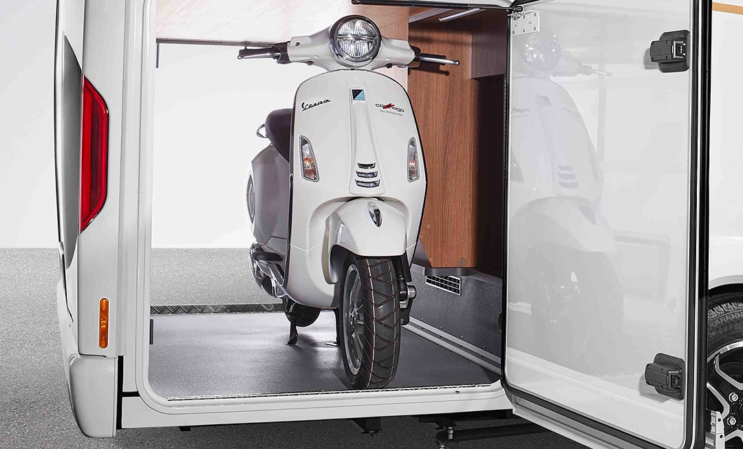 Ein weißer Vespa-Roller, der im Laderaum eines teilintegrierten Carthago c-tourer-Wohnmobils geparkt ist.