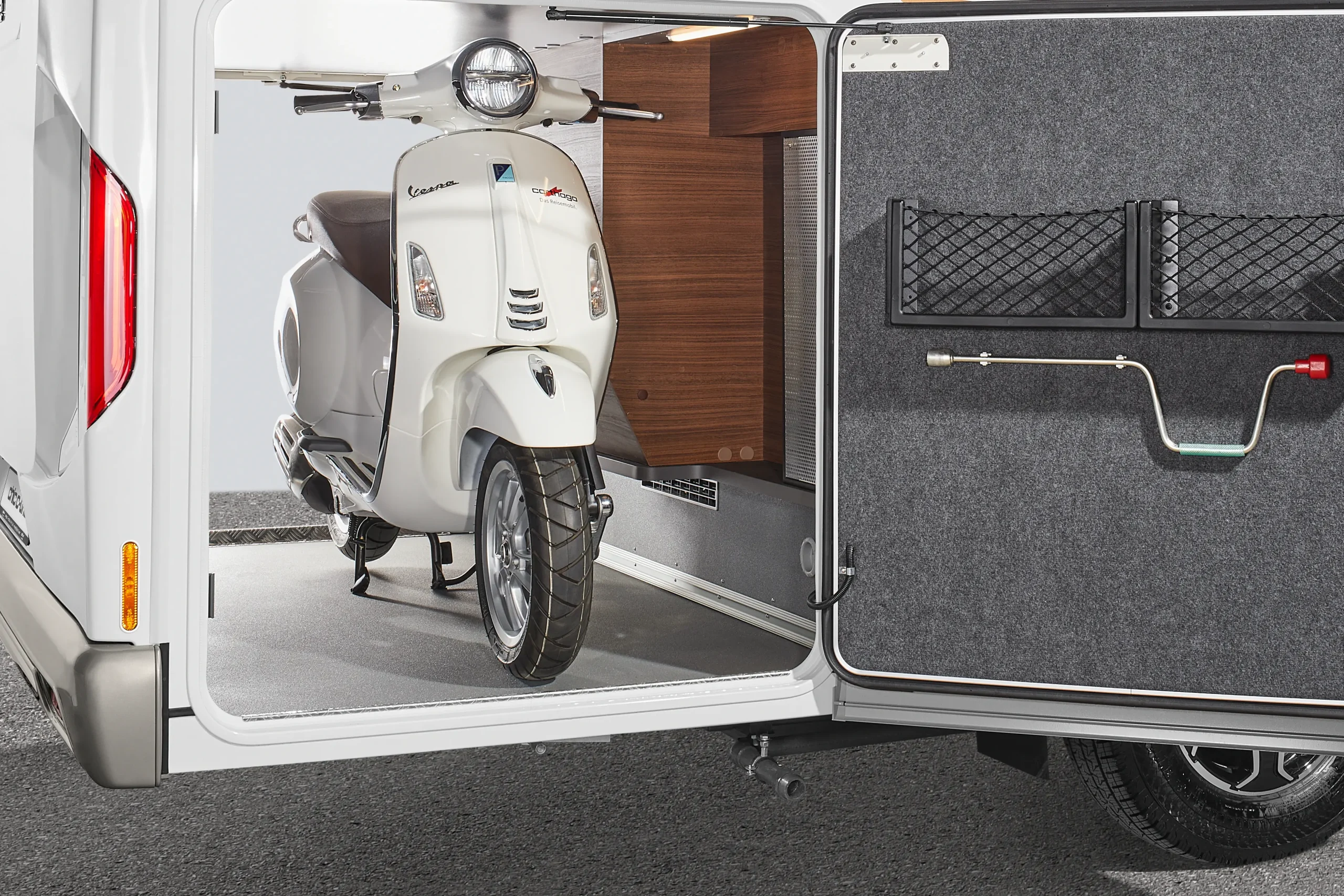 Ein weißer Vespa-Roller, der in der offenen Stauraumgarage eines Carthago Chic C-Line-Wohnmobils geparkt ist.